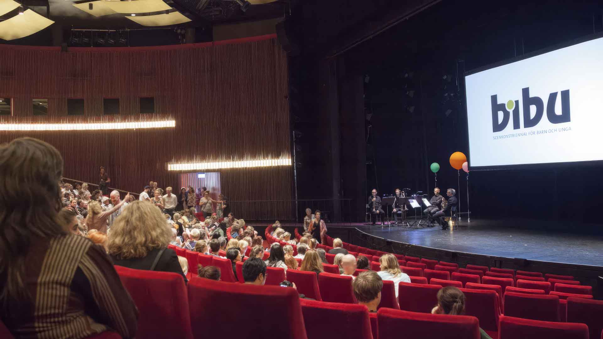 Publik i ett stort auditorium, där några personer sitter på scenen under en skärm med texten "BIBU".