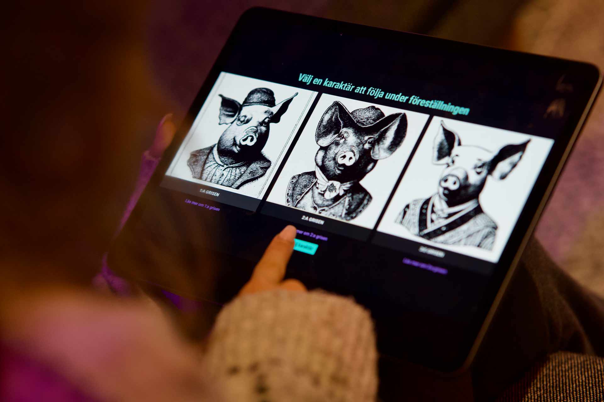 En person i publiken väljer karaktär att följa under en Techformance-föreställning.På surfplattans skärm syns tre tecknade grisar att välja mellan.